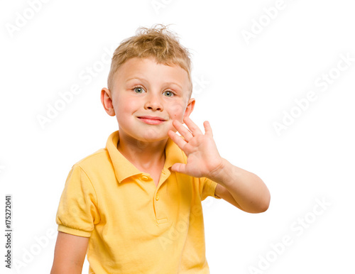 Little boy is greeting someone by waving his hand palm and looking into the distance. Lips smiling face. Blond hair and bright eyes. Dressed in yellow polo shirt. White isolated background.