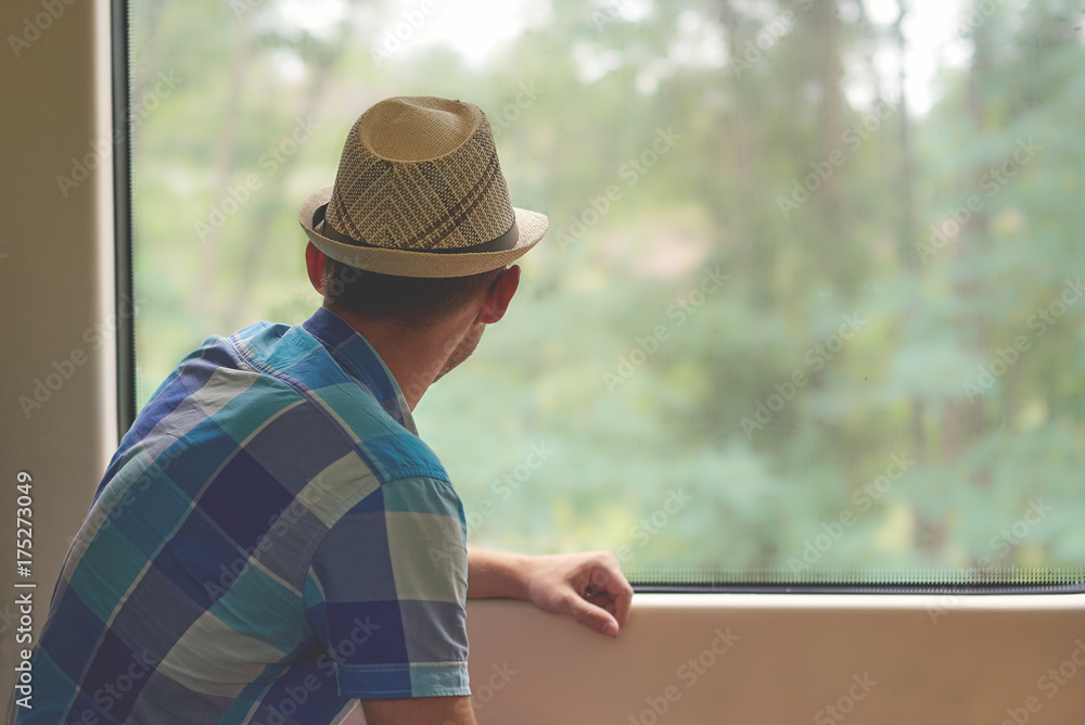 Man in sunhat travelling by train and looking into the window. Back view.