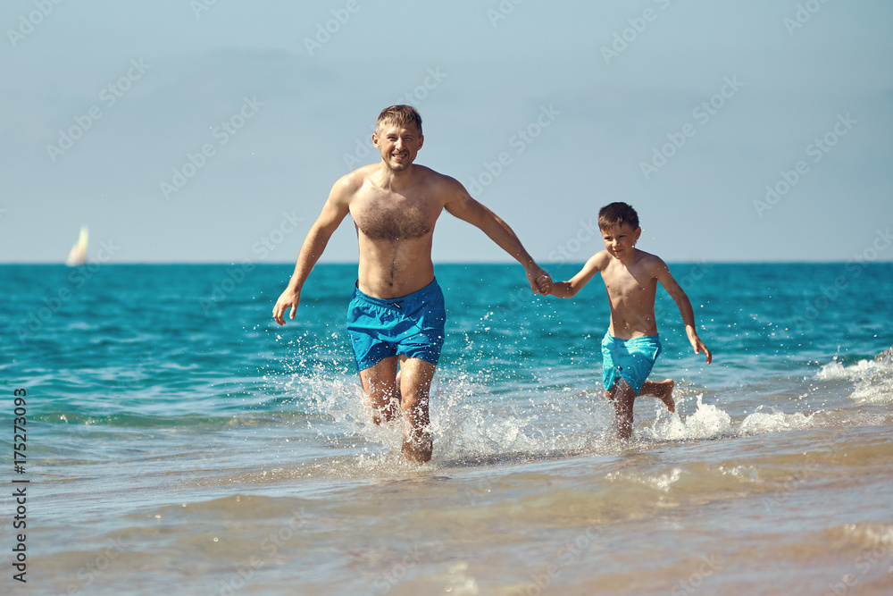 Caucasian dad and son are running in the water along the sea shore. They are holding hands.