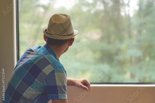 Man in sunhat travelling by train and looking into the window. Back view.
