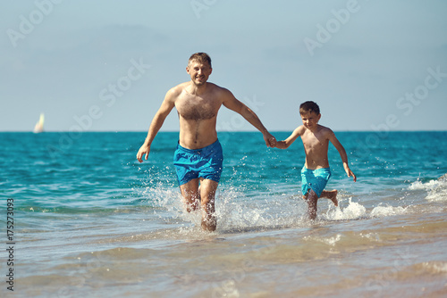 Caucasian dad and son are running in the water along the sea shore. They are holding hands.