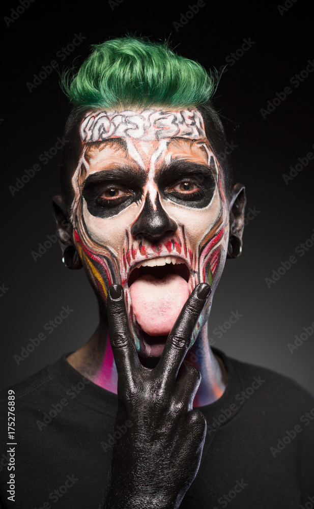 Face art, man with green hair and bright skull makeup showing tongue. Halloween makeup, dead face in color. Face art, man with green hair and bright skull makeup showing tongue. Halloween makeup, dead face in color.