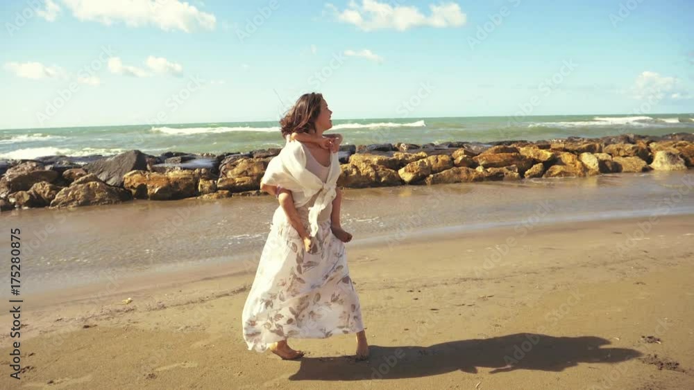 Little girl piggyback on mother singing happy in front of ocean slow motion wide shot Little girl piggyback on mother singing happy in front of ocean slow motion wide shot