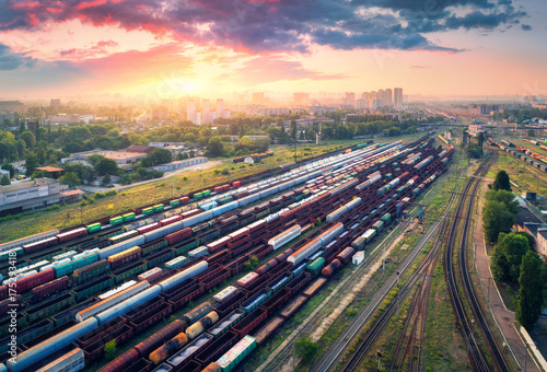 Cargo trains. Aerial view of colorful freight trains. Railway station. Wagons with goods on railroad. Heavy industry. Industrial scene with trains, city buildings and cloudy sky at sunset. Top view 