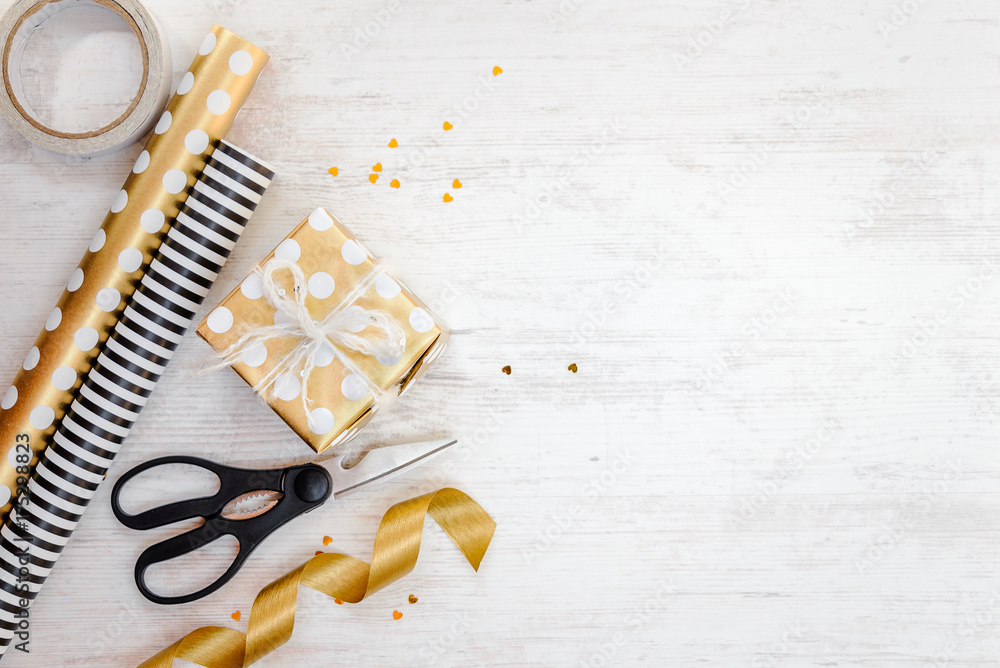 Gift box wrapped in golden dotted paper and wrapping materials on a white wood old background. Empty space.