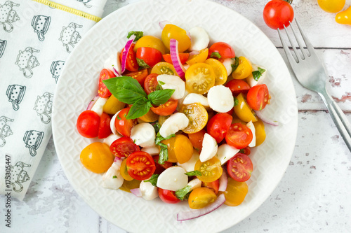 Homemade caprese salad with fresh yellow and red cherry tomatoes, mozzarella cheese, red onion and basil on kitchen table, top view.