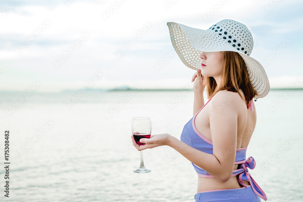 Fototapeta premium Sexy girl in a swimsuit walking on the beach and holding a glass of wine in hand, on a beautiful sunny day on the weekends.