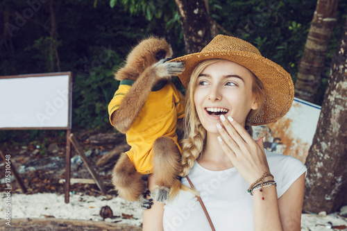 Close up of beautiful young woman with small dress up monkey on her shoulder.
She is smiling and enjoying time with cute little baby monkey on the lonely beach with palm trees. 