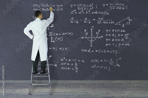 Scientist of genius standing on a ladder writing complex mathematical formulas with white chalk on large blackboard.