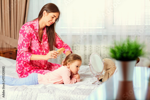 Mother combs hair to daughter sitting on the bed in the room.