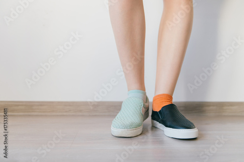 Female naked legs standing on wooden floor in front of white wall. Women`s feet in different leather shoes. Missmatched socks. Odd unusual weird unrecognizable bizarre kinky girl. Soft taned skin.