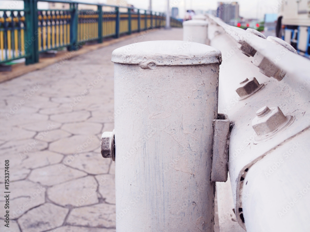 Close up white painted short steel round column and wall, with stone tile footpath floor, traffic car on paved road, and bridge fence, under warm evening sunlight background Close up white painted short steel round column and wall, with stone tile footpath floor, traffic car on paved road, and bridge fence, under warm evening sunlight background