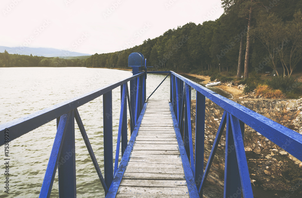 Obraz premium Mole (pier) on the scandinavian lake with motorboat on side and misty hills. Blue wooden bridge in forest in autumn time with blue lake. Lake for fishing with pier and powerboat. Foggy lake with hills