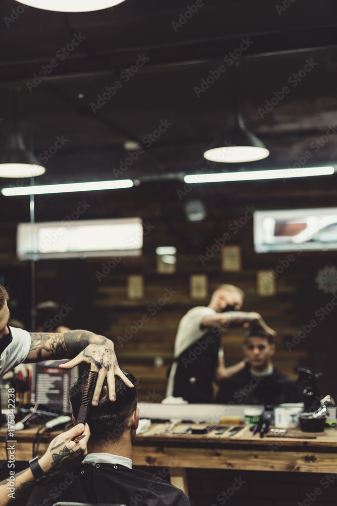 Anonymous stylish barber with tattoos cutting hair of male client in chair.