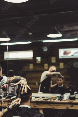 Anonymous stylish barber with tattoos cutting hair of male client in chair.