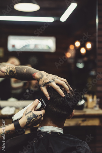 Crop tattooed stylist concentrated on shaving man with machine doing hairstyle.