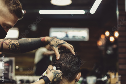 Fotografie Crop tattooed stylist concentrated on shaving man with machine doing hairstyle