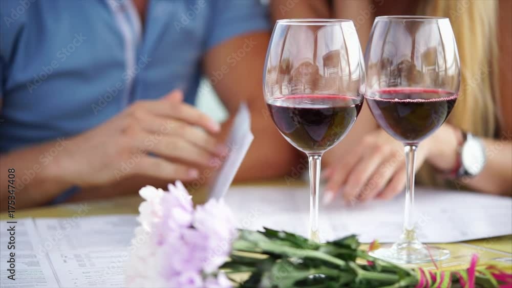Close up shot of glass glasses of red wine, in the background sit a man and woman and view the menu, a couple in love is in a restaurant and choose the main dish Close up shot of glass glasses of red wine, in the background sit a man and woman and view the menu, a couple in love is in a restaurant and choose the main dish