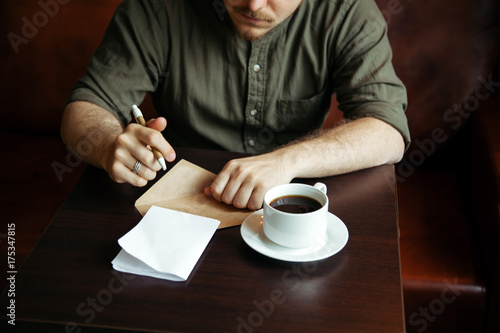 Young serious fashionable man sitting alone and writing a letter in loft-styled cafe. Cup of black coffe is standing on his table. Former factory building  natural daylight.