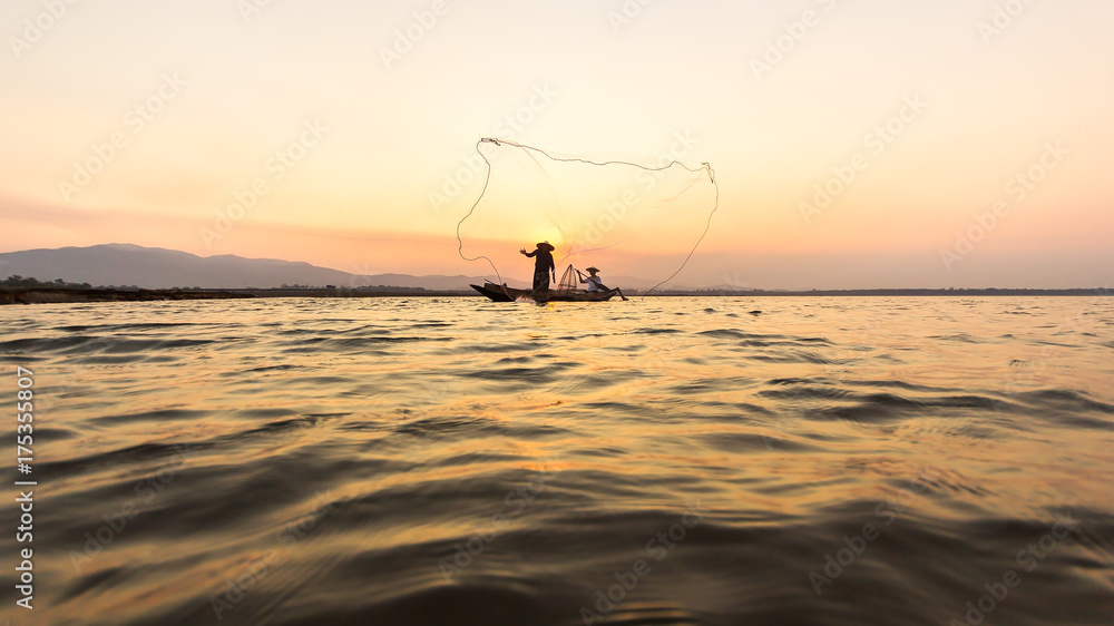 Silhouette of asian fisherman on wooden boat ,fisherman in action throwing a net for catching freshwater fish in nature river, traditional fishermen at the sunset .