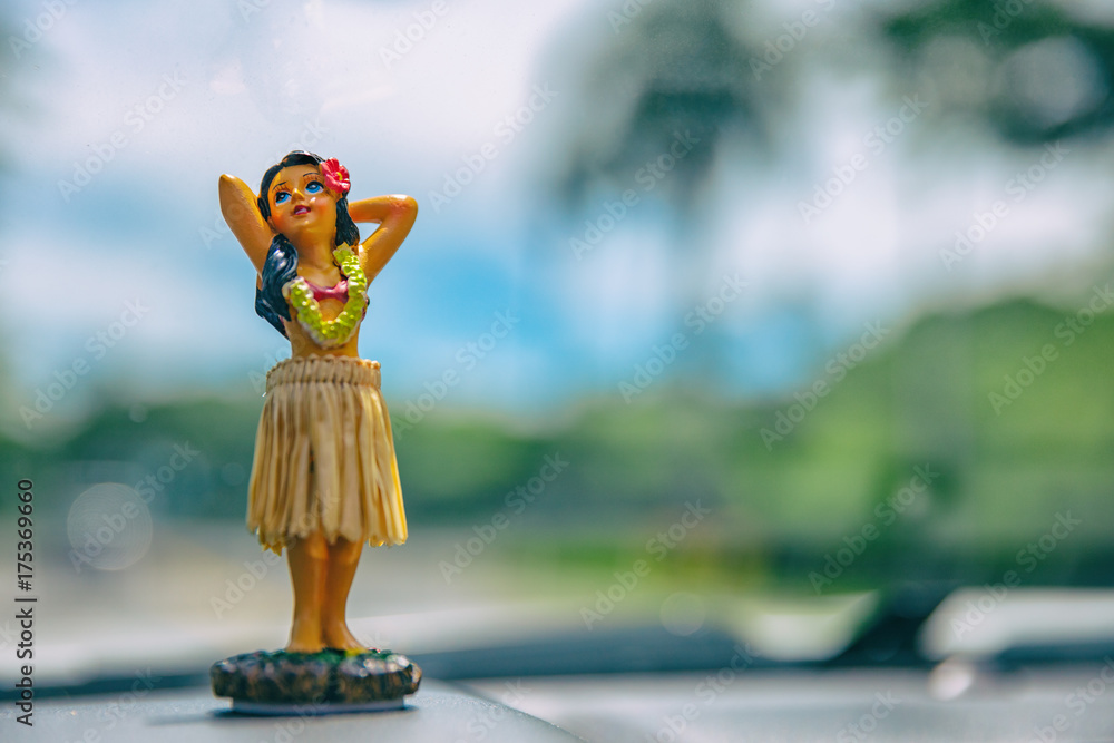 Naklejka premium Hula dancer doll on Hawaii car road trip travel vacation. Aloha mini girl doll dancing on the dashboard in tropical nature landscape. Tourism and Hawaiian holiday freedom concept.