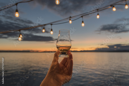 Fototapete Old fashioned whiskey in hand being held up over the sea at sunset under hanging deck lights