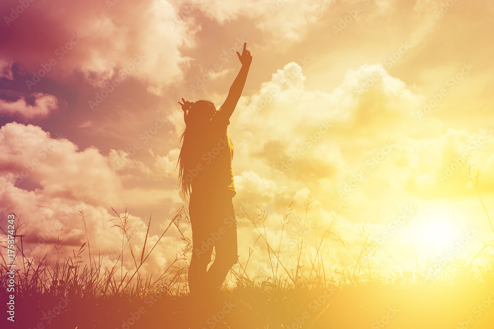 Silhouette young woman with raised hands standing on meadow on sunset, light effect