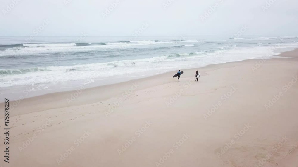 Young couple of friendly surfers wearing wetsuit holding surfboards, walking by the sea on a morning surfing session. Young couple of friendly surfers wearing wetsuit holding surfboards, walking by the sea on a morning surfing session.