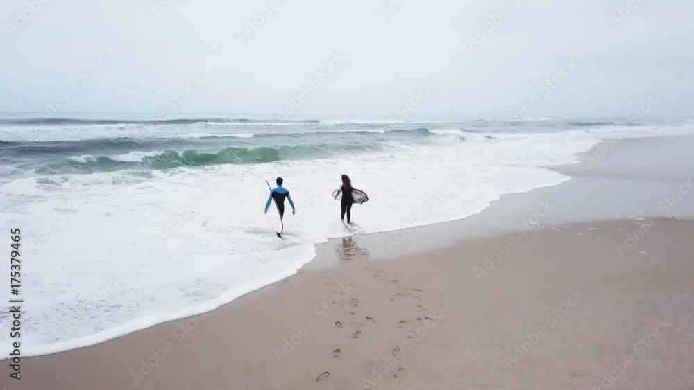 Young couple of friendly surfers wearing wetsuit holding surfboards, walking by the sea on a morning surfing session. Young couple of friendly surfers wearing wetsuit holding surfboards, walking by the sea on a morning surfing session.