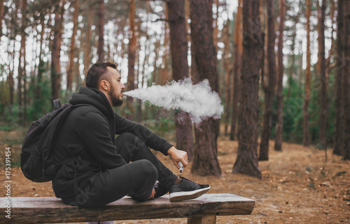 brutal bearded young man having rest in forest and vaping vape device. Lifestyle. Vape concept . Rest in forest photo