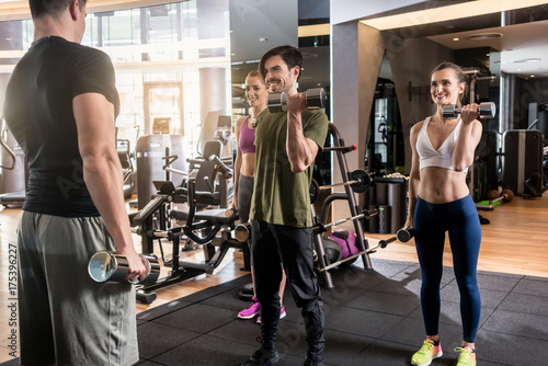 Εκτύπωση καμβά Low-angle view of three young people smiling while alternating dumbbell bicep cu