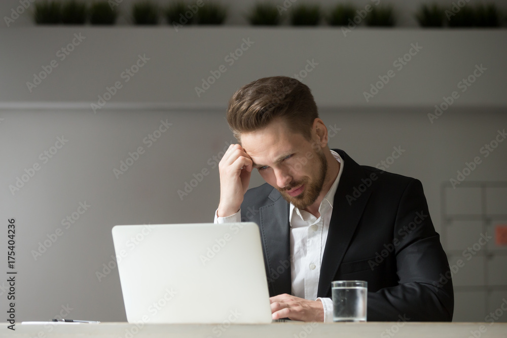 Perplexed, puzzled handsome businessman looking at laptop screen at workplace in office. Young CEO or project manager is not sure whether he should agree to the deal, sign contract, buy stock shares. Perplexed, puzzled handsome businessman looking at laptop screen at workplace in office. Young CEO or project manager is not sure whether he should agree to the deal, sign contract, buy stock shares.