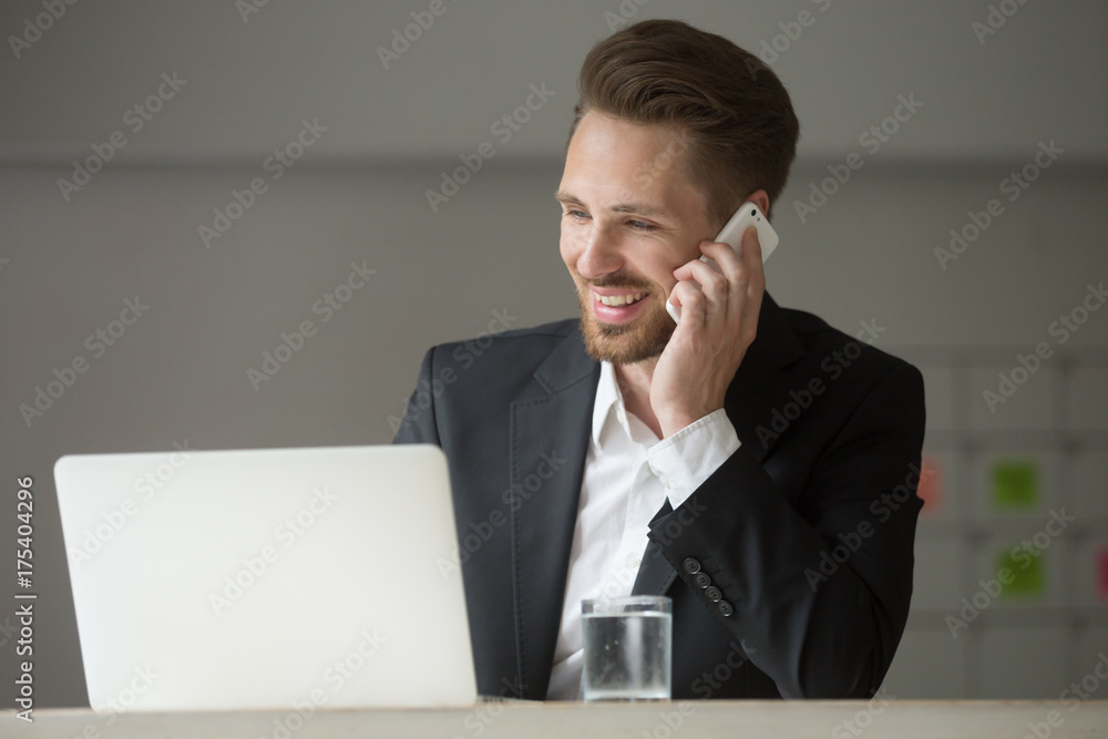 Happy smiling young businessman in office at work desk in front of laptop talking on cellphone. Successful deal discussion, planning future meetings, business negotiations, client consulting concept. Happy smiling young businessman in office at work desk in front of laptop talking on cellphone. Successful deal discussion, planning future meetings, business negotiations, client consulting concept.