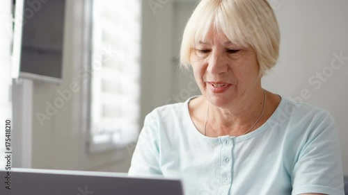 Senior woman sitting at home with laptop. Using computer talking via messenger app Skype. Smiling waving hand in greeting. Technology and communications use in active modern lifestyle of older people