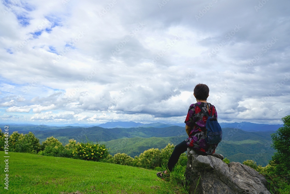 Woman sitting on the rock and looking at the view of beautiful mountains with cloudy sky at  Pha Chu, Sri Nan National Park, Nan, Thailand
