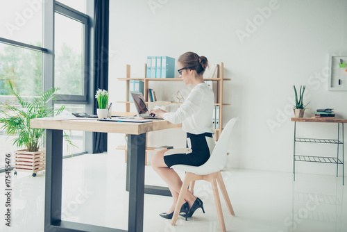 Full length side shot of gorgeous business lady, sitting at her light modern work station, checking reports on her digital device, in trendy specs, with hairdo. Development, authority, feminity