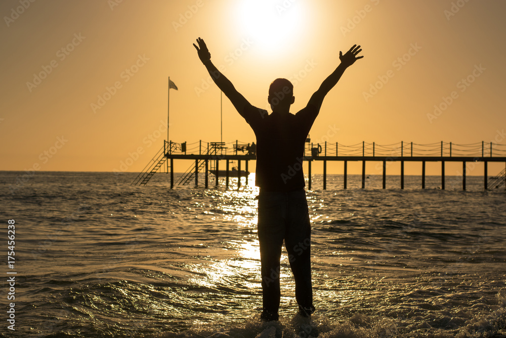Fototapeta premium Man standing on the beach, raising his hands above his head at the sunset