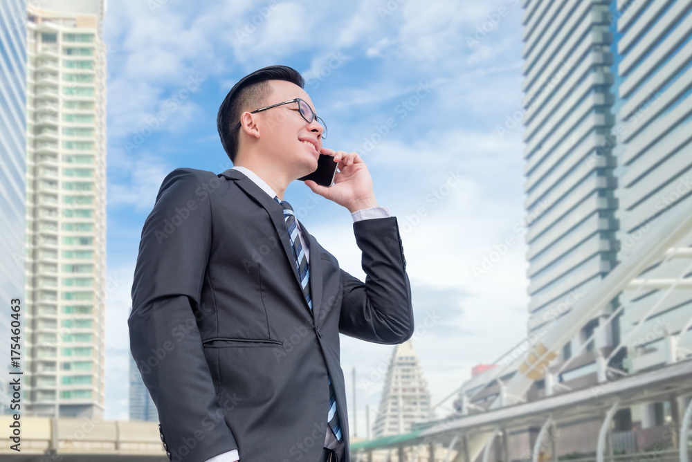 Young asian businessman wearing black suit talking via smart phone with modern building in background Young asian businessman wearing black suit talking via smart phone with modern building in background