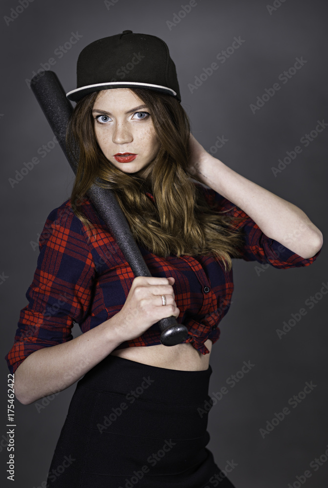 Half body indoor portrait of young attractive woman dressed in baseball cap and red-blue plaid shirt holding pearl-grey bat in hand lay on shoulder Half body indoor portrait of young attractive woman dressed in baseball cap and red-blue plaid shirt holding pearl-grey bat in hand lay on shoulder