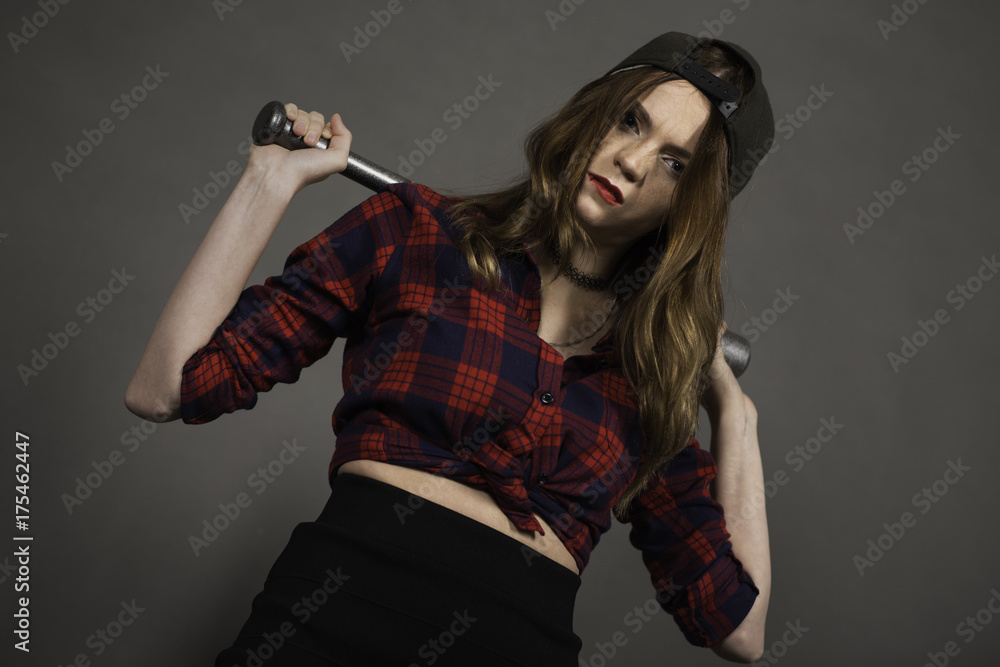 Indoor portrait of young attractive woman dressed in baseball cap and red-blue plaid shirt holding pearl-grey bat in hands on shies back neck and looking to camera. Grey background Indoor portrait of young attractive woman dressed in baseball cap and red-blue plaid shirt holding pearl-grey bat in hands on shies back neck and looking to camera. Grey background