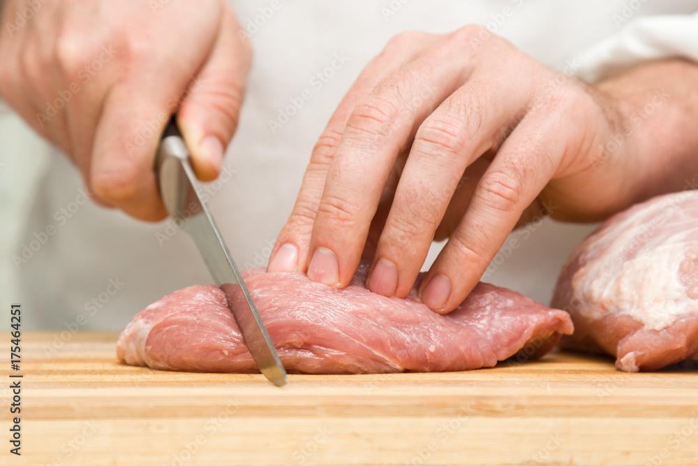 Chef's hands with a knife cutting a meat on the wooden board in the kitchen. Preparation for cooking. Food concept.