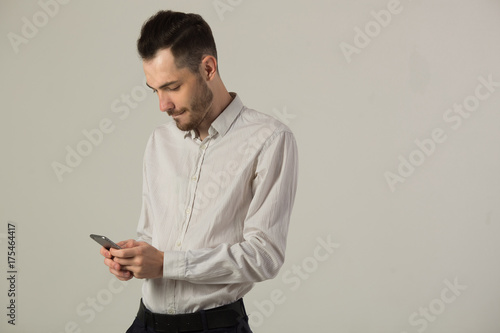 young brunette man in white shirt with unshaven face on background of light wall with phone in hand © Alexandr