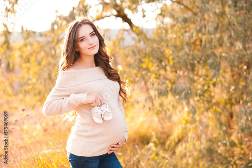 Happy pregnant woman 24-25 year old holding baby shoes outdoors. Wearing beige knitted sweater over autumn background. Looking at camera. Maternity. Motherhood.