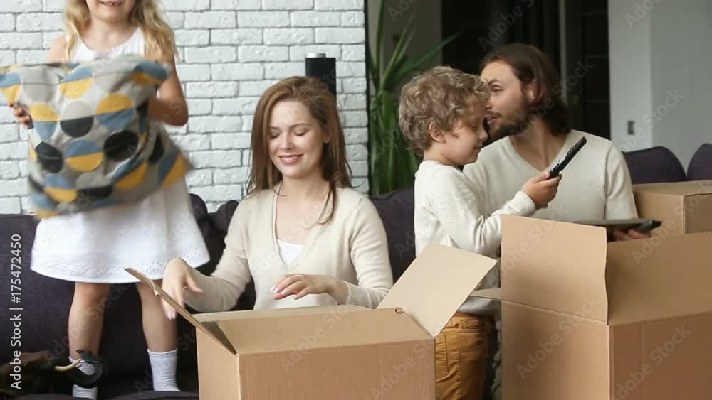 Happy excited family with two children unpacking boxes moving into new home concept, small kids helping parents with belongings in cardboards putting cushion on sofa in living room after relocation Happy excited family with two children unpacking boxes moving into new home concept, small kids helping parents with belongings in cardboards putting cushion on sofa in living room after relocation