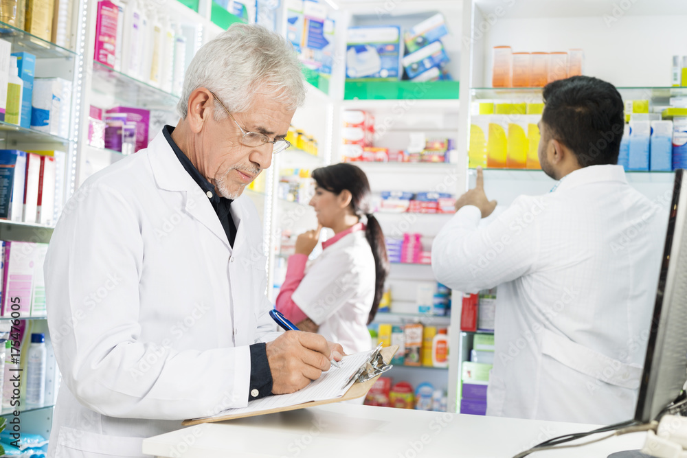 Chemist Writing On Clipboard While Coworkers Working In Pharmacy Chemist Writing On Clipboard While Coworkers Working In Pharmacy