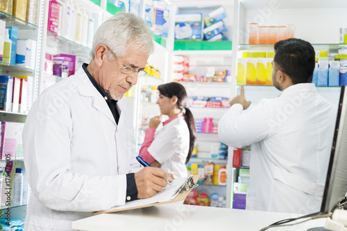 Chemist Writing On Clipboard While Coworkers Working In Pharmacy