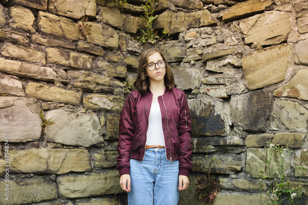 young girl with glasses posing near a wall of stones
