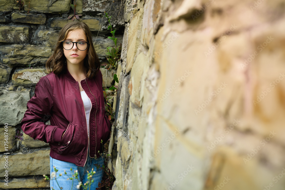 young girl with glasses posing near a wall of stones