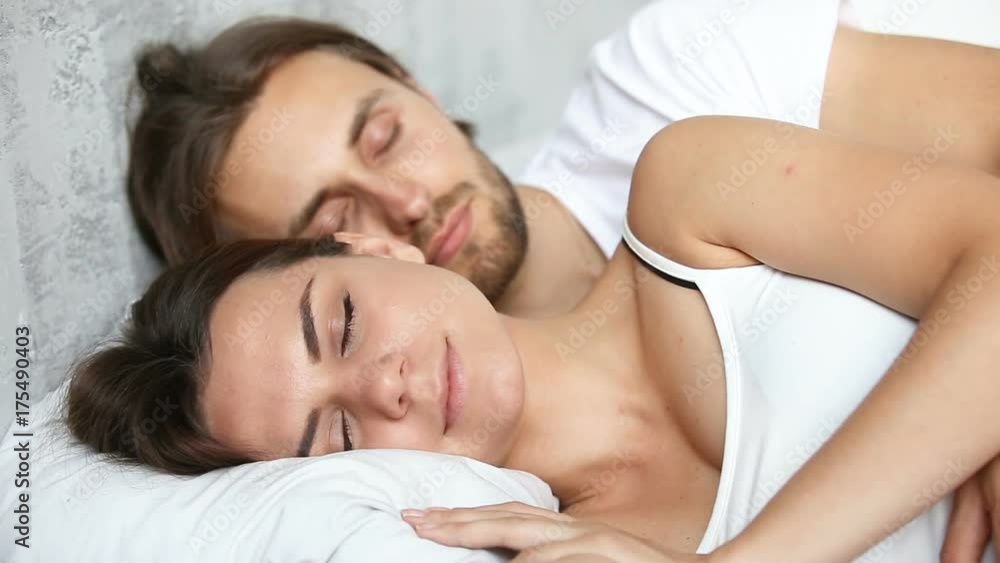 Young couple sleeping in bed, loving husband embracing wife lying on white linen with eyes closed enjoying smiling in light nap in the morning before waking up, happy woman and man resting together Young couple sleeping in bed, loving husband embracing wife lying on white linen with eyes closed enjoying smiling in light nap in the morning before waking up, happy woman and man resting together