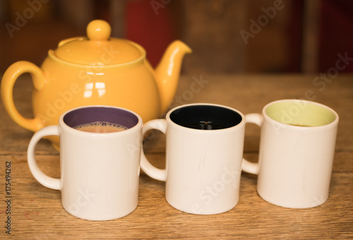 Three cups of tea and a teapot are on a wooden oak table. One cup of tea with milk, while others have black tea without milk.